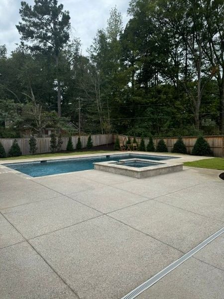 Concrete patio with a pool and spa, surrounded by trees and a wooden fence.