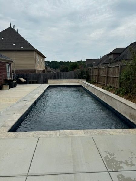 Rectangular backyard swimming pool, concrete deck, surrounded by fence and homes, cloudy sky.