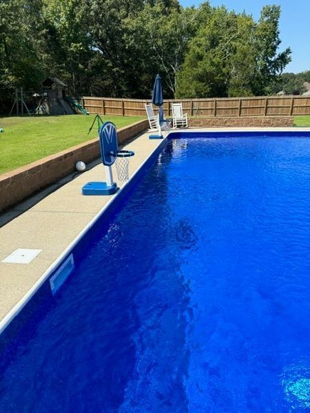 Blue pool with basketball hoop and two white chairs, grassy yard in the background.