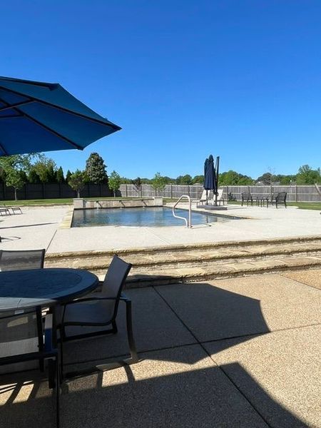 Poolside scene with pool, patio, blue umbrella, table, chairs, and sunny sky.