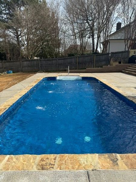 Rectangular outdoor pool with blue water, surrounded by stone tiles. Trees and a house in the background.