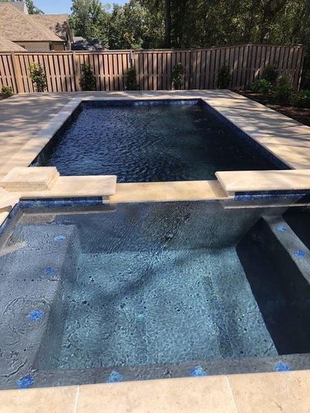 Rectangular pool with attached spa, surrounded by stone patio. Wooden fence in background.