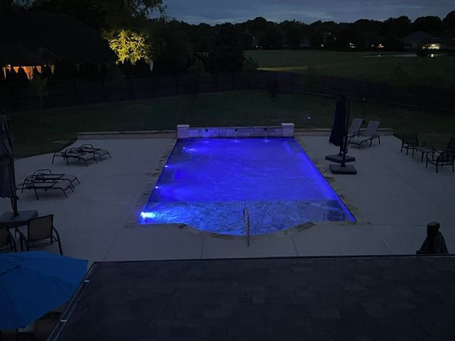 A nighttime view of a rectangular pool with blue lights, surrounded by patio furniture and a dark lawn.