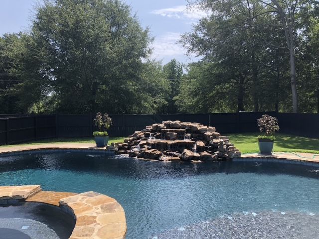 Pool with waterfall feature and surrounding trees. Blue water, stone, and green foliage.