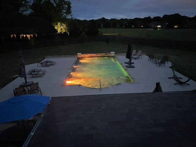 Night view of a rectangular pool with colorful underwater lights, surrounded by a concrete patio and lounge chairs.