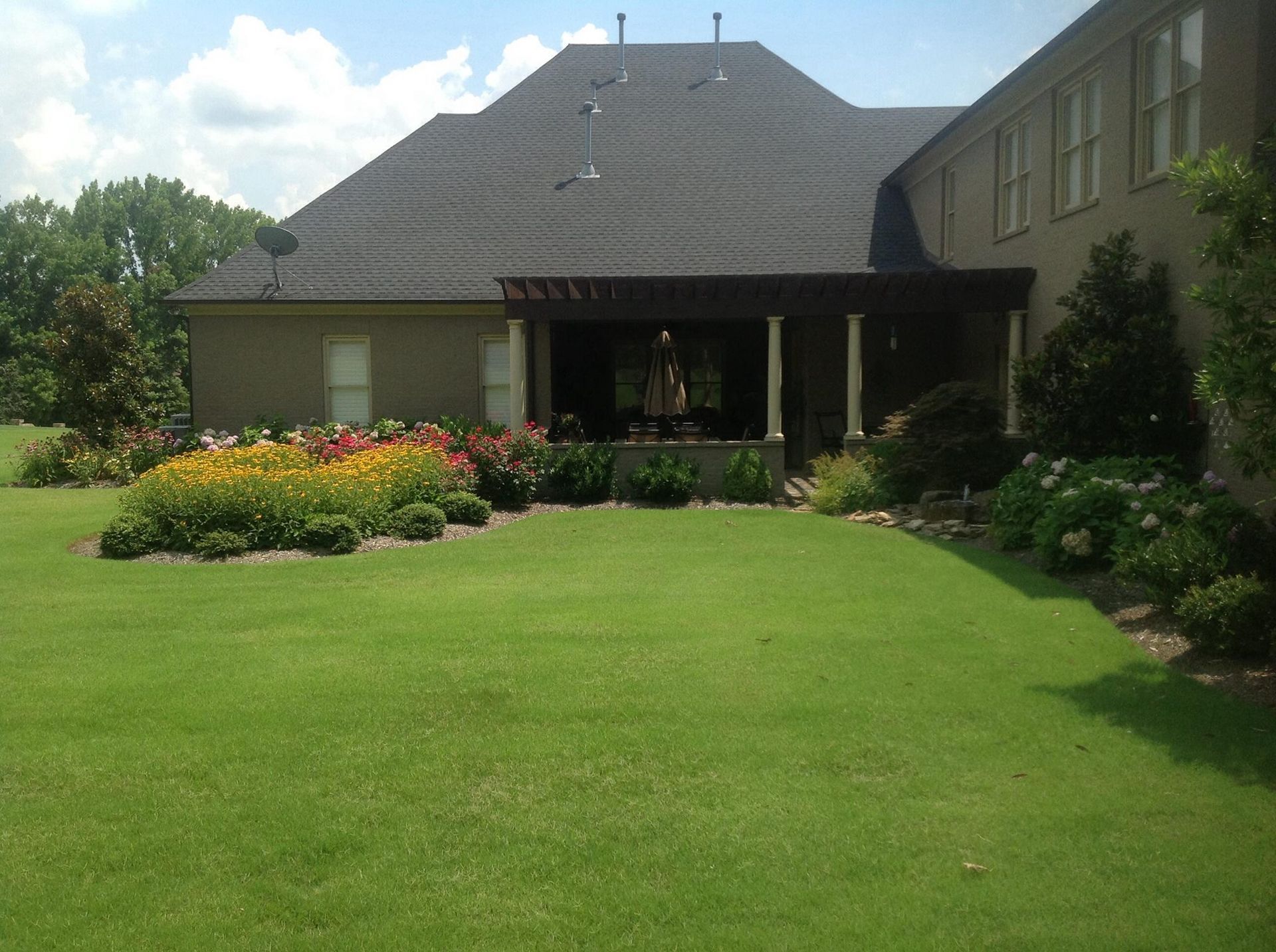 Backyard with lawn, flowerbeds, and covered patio attached to a house with a dark roof.