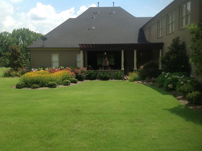Backyard with lawn, flowerbeds, and covered patio attached to a house with a dark roof.