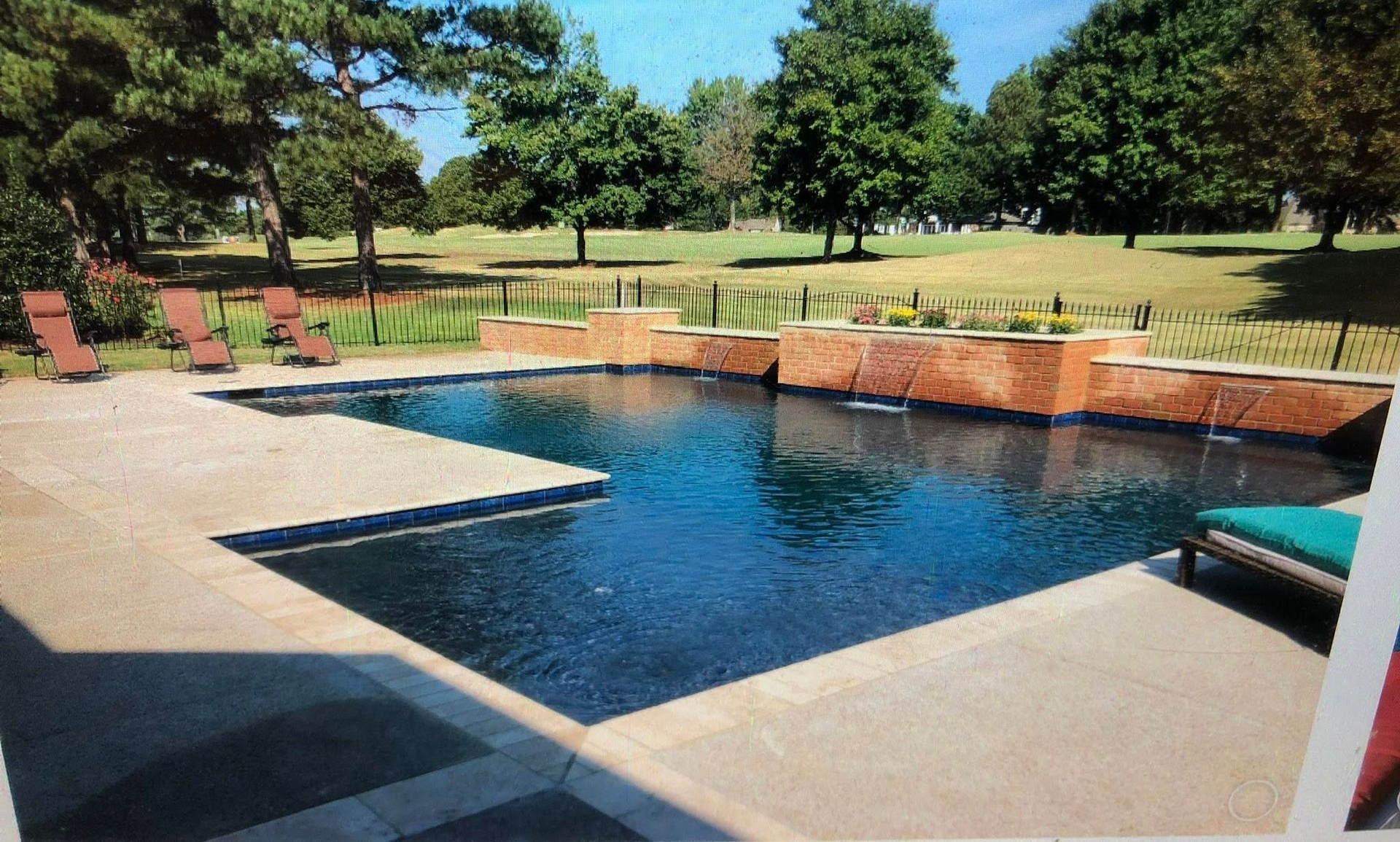 Pool with dark blue water and stone surround, patio with lounge chairs. Trees and grass in background.