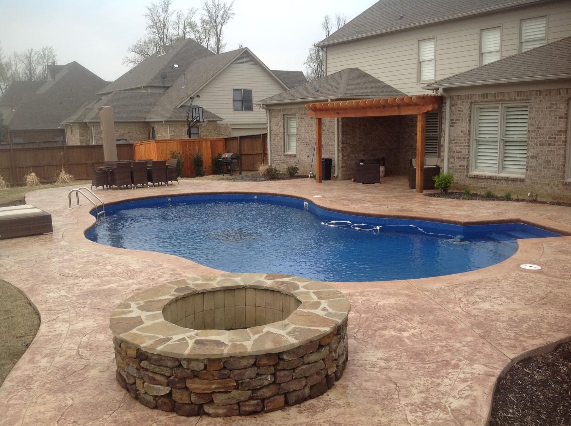 Backyard pool with a stone fire pit and seating area, surrounded by a stamped concrete patio and a house.
