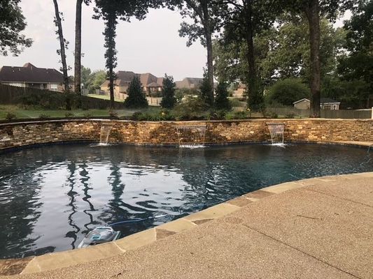 Pool with waterfalls and stone wall; trees and houses in the background.