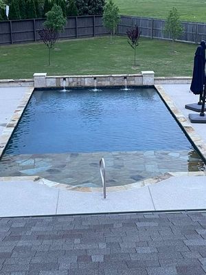 Rectangular pool with cascading fountains, stone steps, and surrounding patio. Green lawn and trees in background.