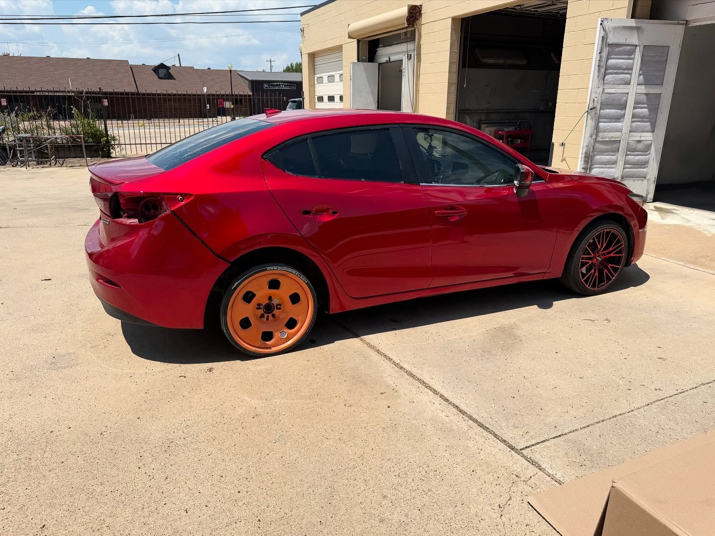 Red sedan parked outside a building, orange wheels, sunny day.