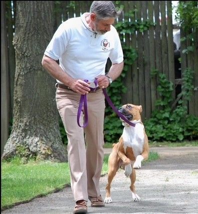 Man training a dog on a leash; woman watches in the background. Outdoors, on a path.