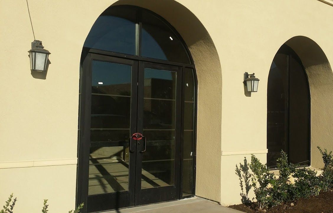 Arched glass double doors with a red handle on a beige stucco building, flanked by wall lanterns