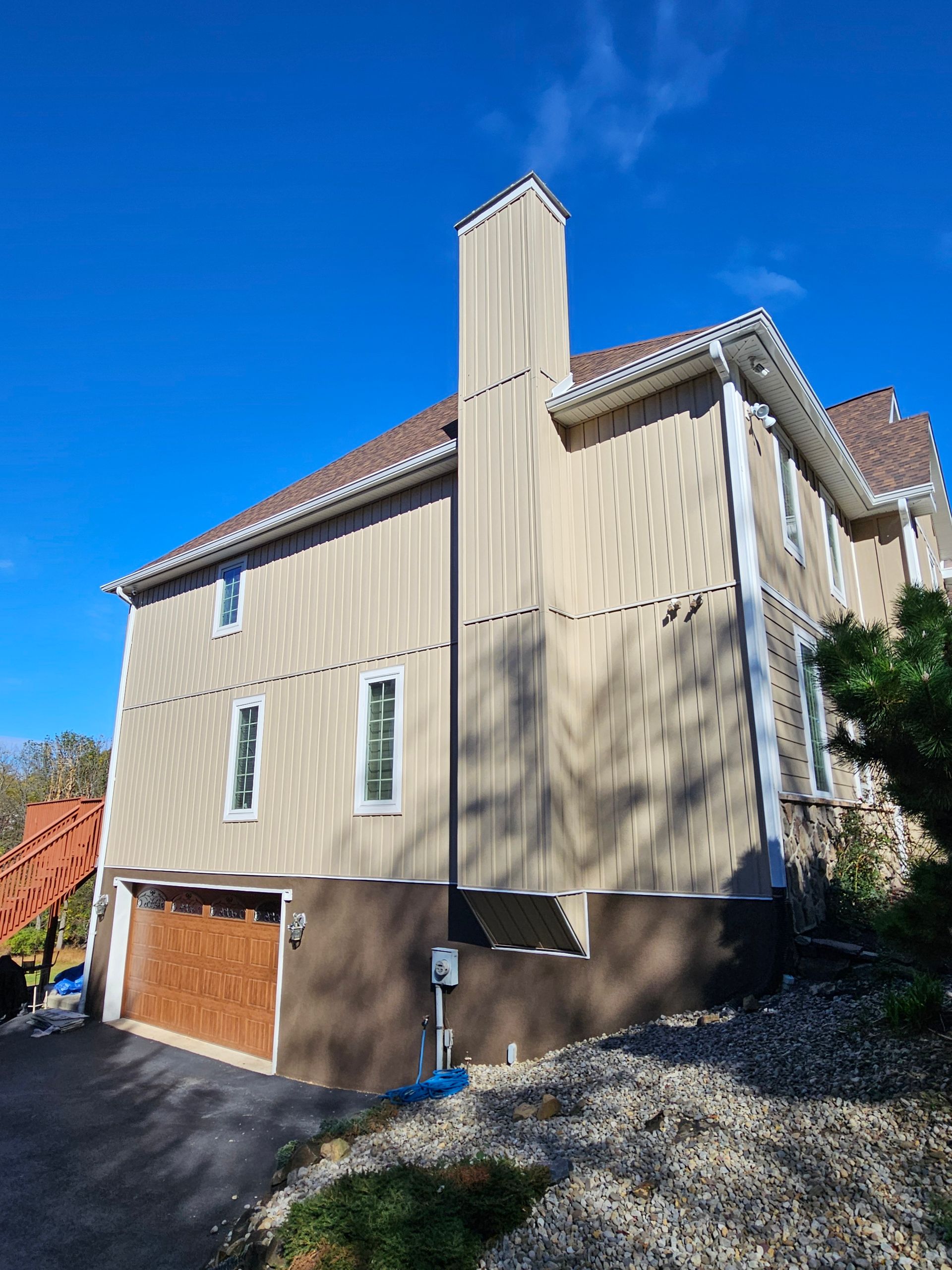 Tan house with chimney and brown garage door against a blue sky, on a slight hill.