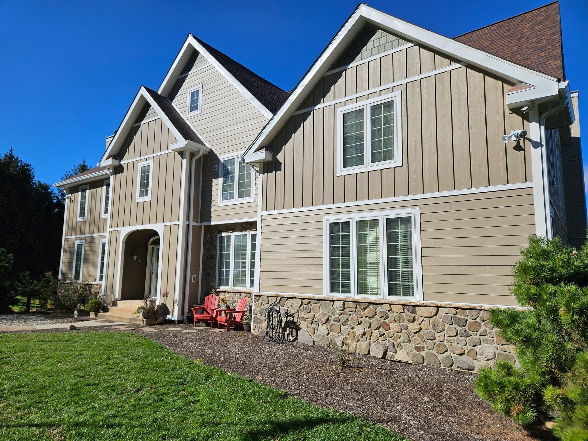 Two-story beige house with stone base, white trim, and brown roof under a blue sky.