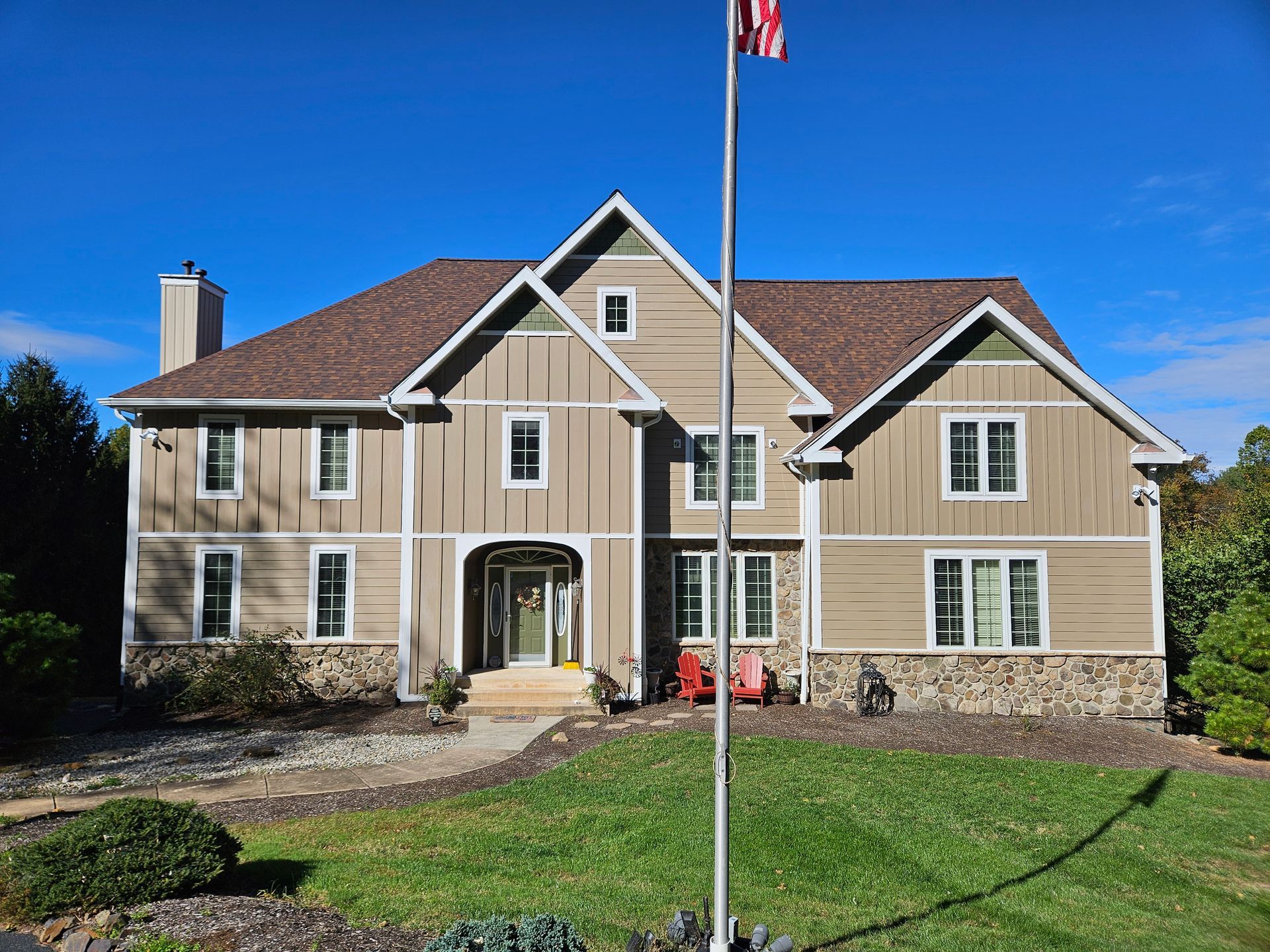 Two-story house with tan siding, stone accents, and brown roof under a blue sky, with a flagpole in the yard.