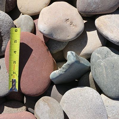 Pile of smooth river rocks, including a reddish-brown stone near a yellow tape measure showing size.