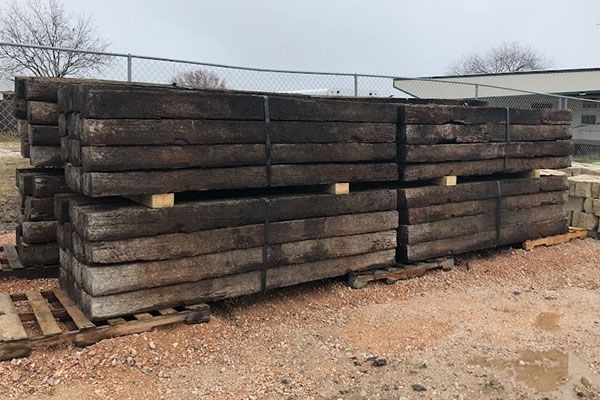 Stacks of weathered, dark-brown railway ties on wooden pallets, outdoors with a fence and overcast sky.