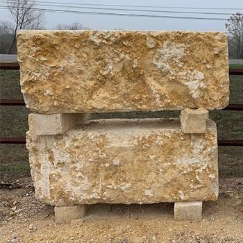 Two rough, light-brown stone blocks stacked, one on top of the other, supported by four smaller blocks. Outdoors.