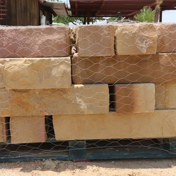 Stacked rectangular sandstone blocks wrapped in wire mesh, varying tan and reddish-brown colors.