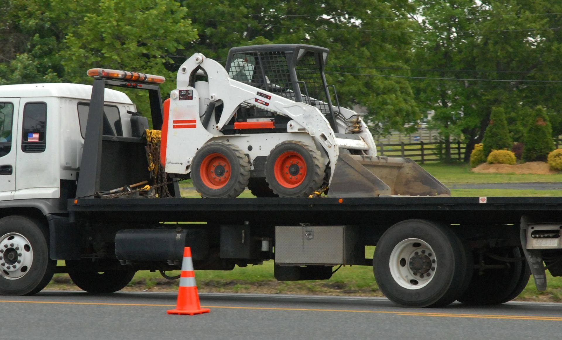 White Bobcat skid-steer loader on a flatbed tow truck with an orange traffic cone on the road.