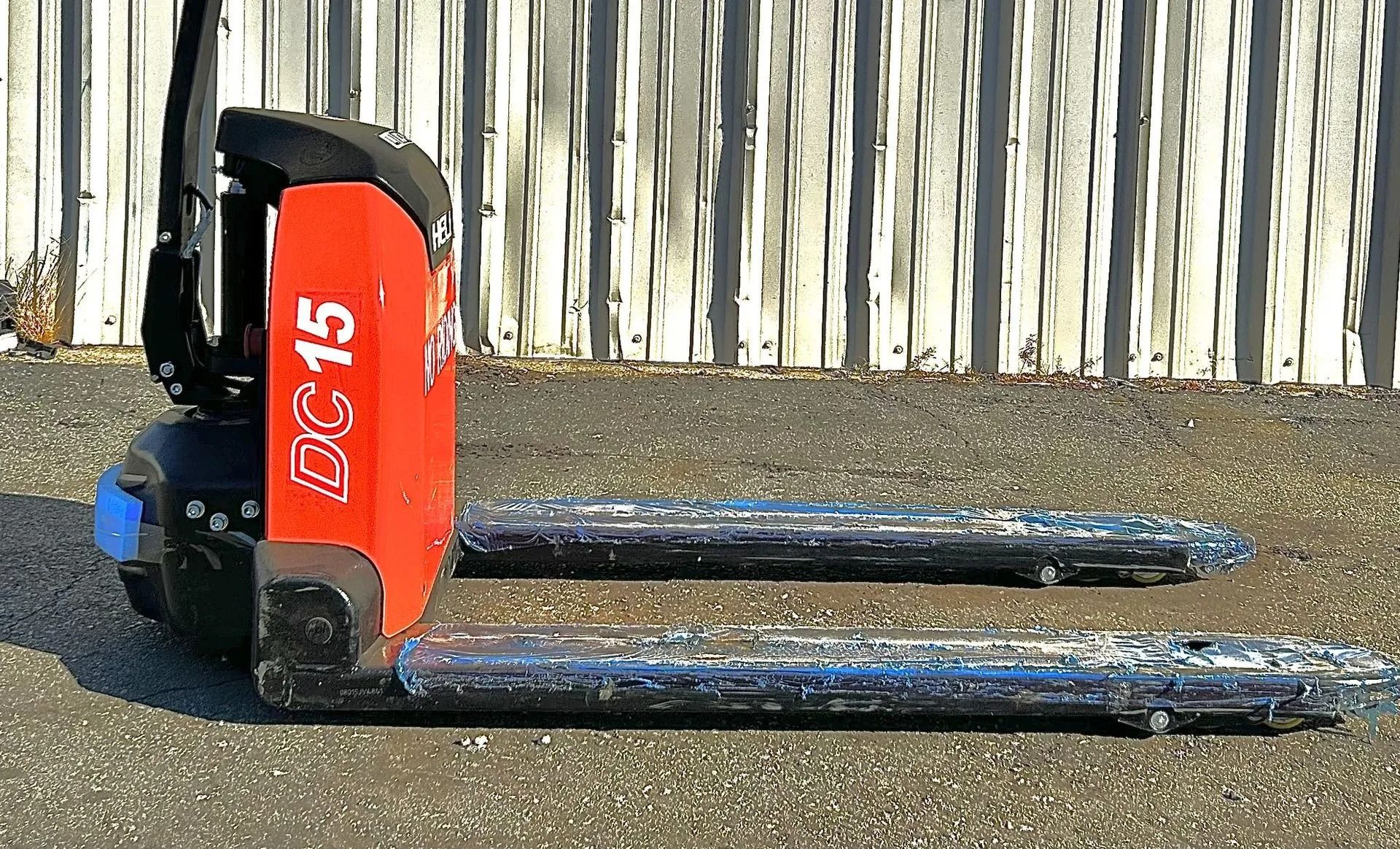 Red and black electric pallet jack with extended forks, parked outside against a weathered building.
