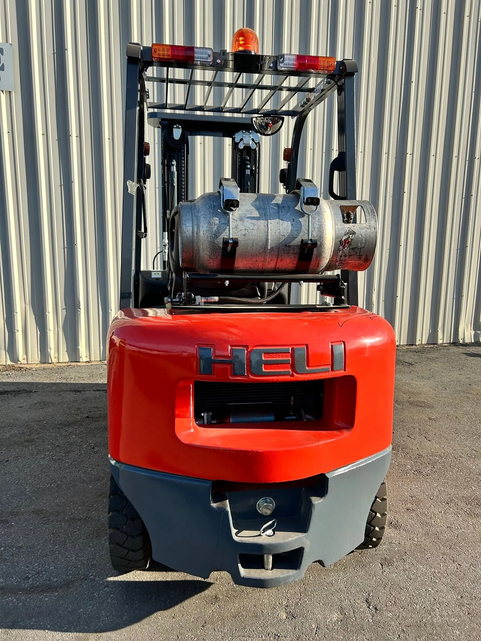 Red and gray HELI forklift with a propane tank, against a corrugated metal wall.