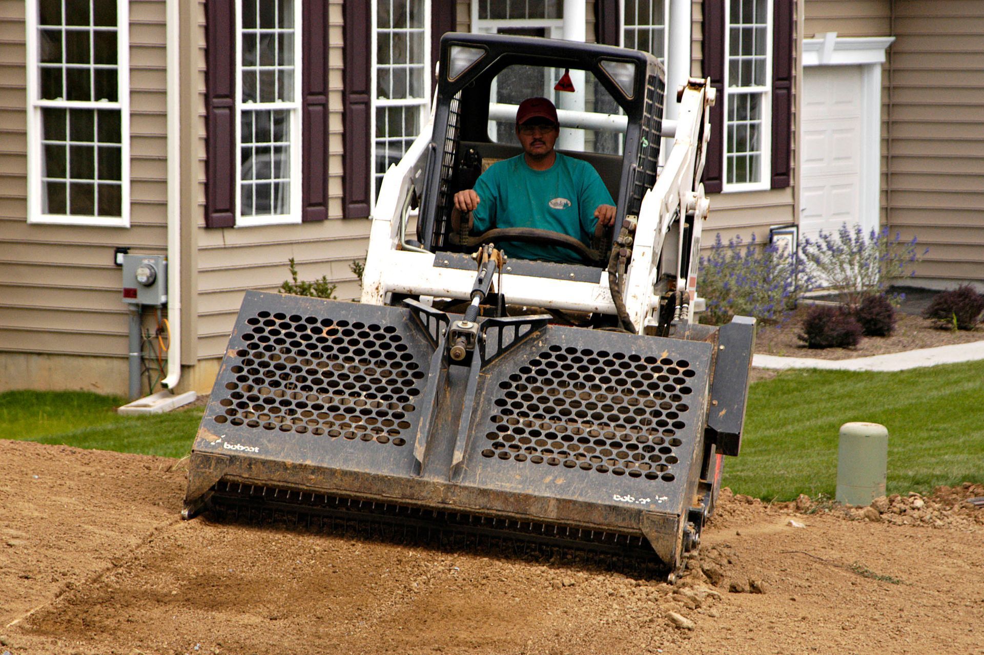 Bobcat skid-steer with operator on dirt, in front of a house, landscaping the area.