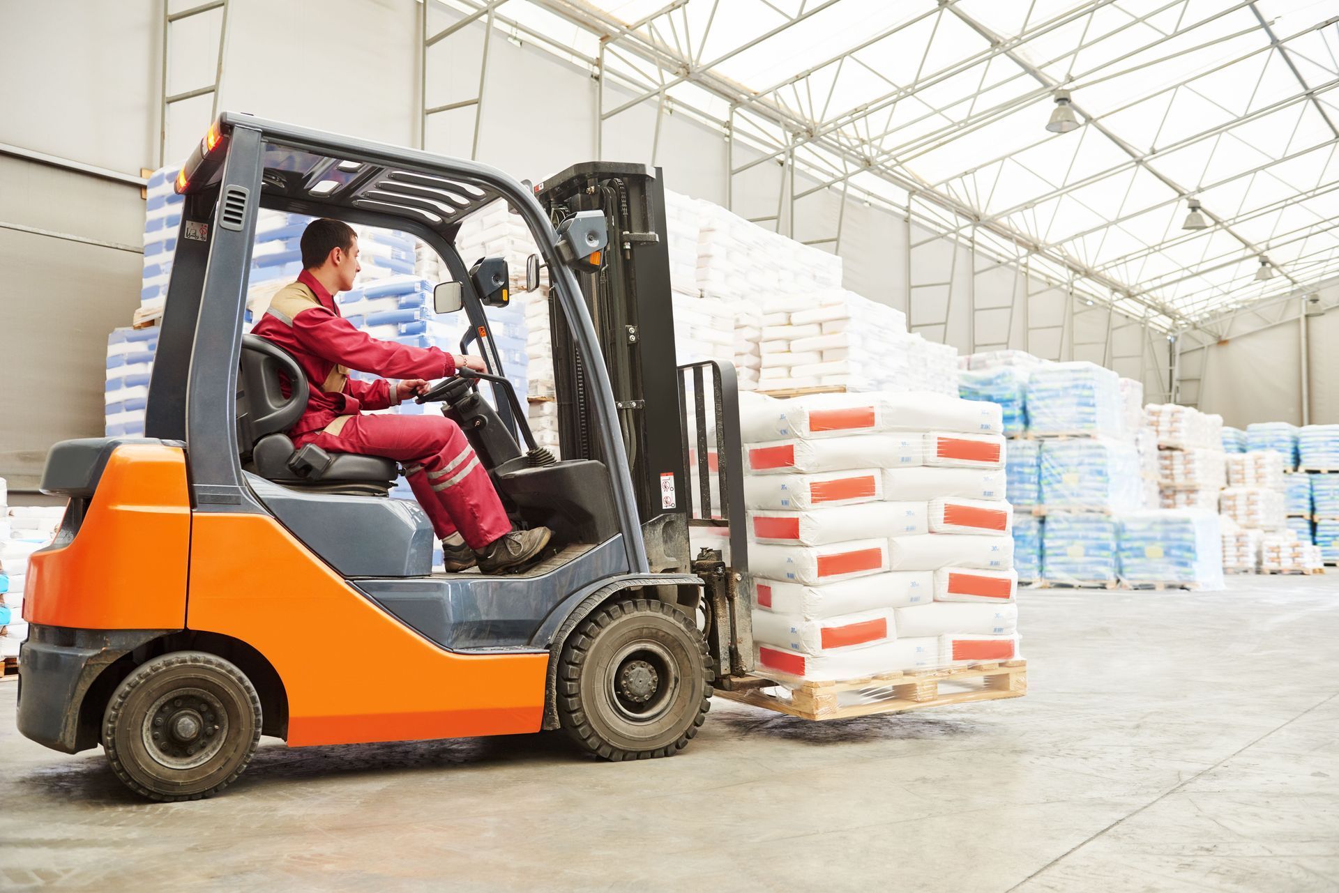 Forklift operator moving a pallet of white bags in a warehouse.