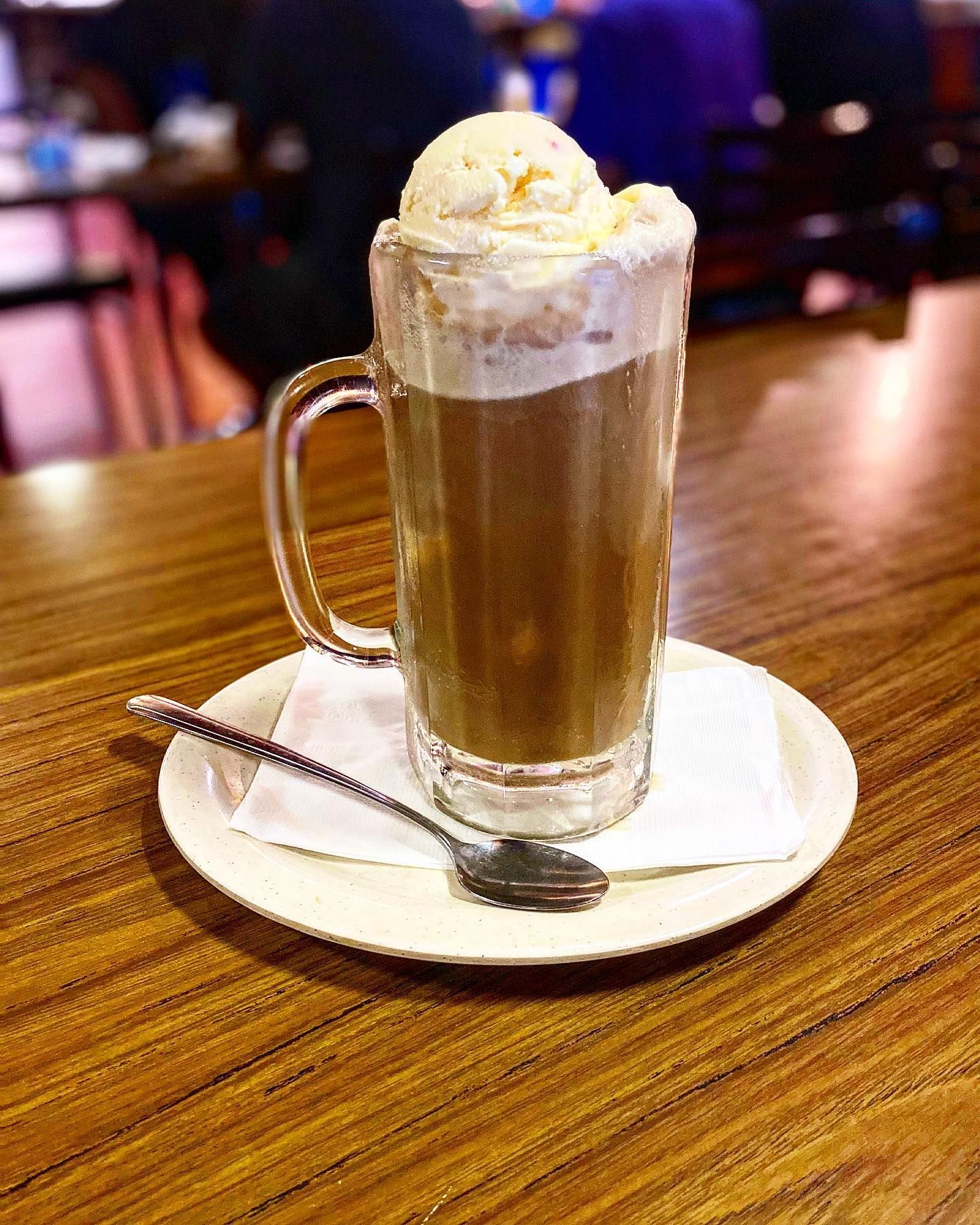 A glass of coffee with ice cream on top is on a plate on a wooden table.
