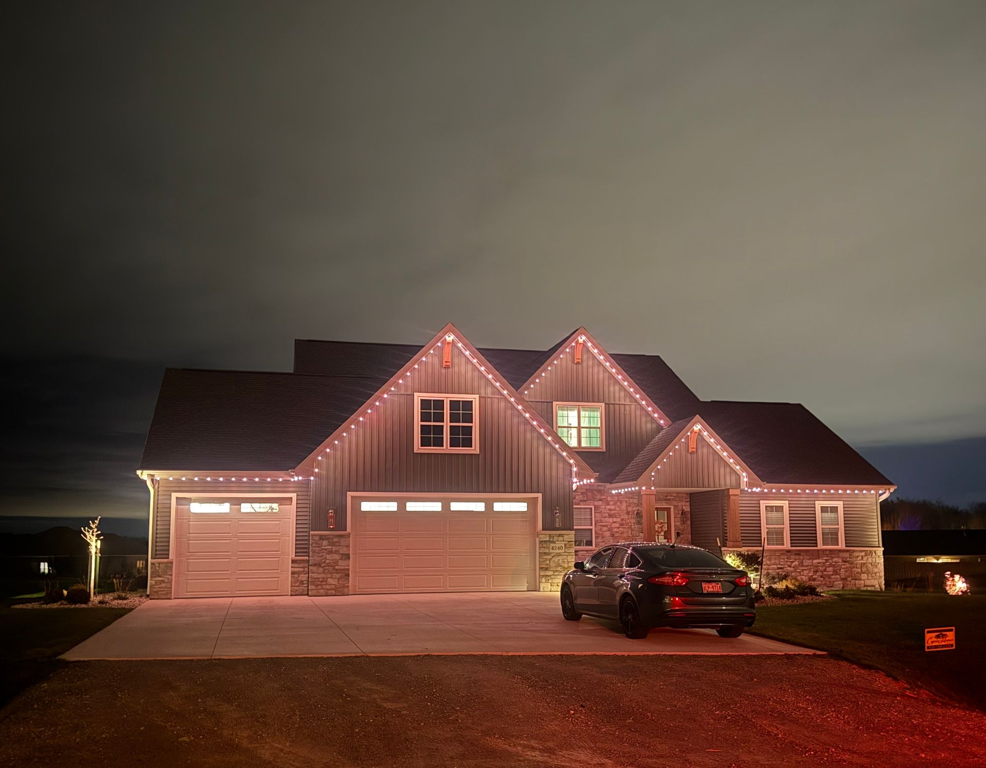 House decorated with red, white, and blue Christmas lights at night. Garage and porch light up the yard.
