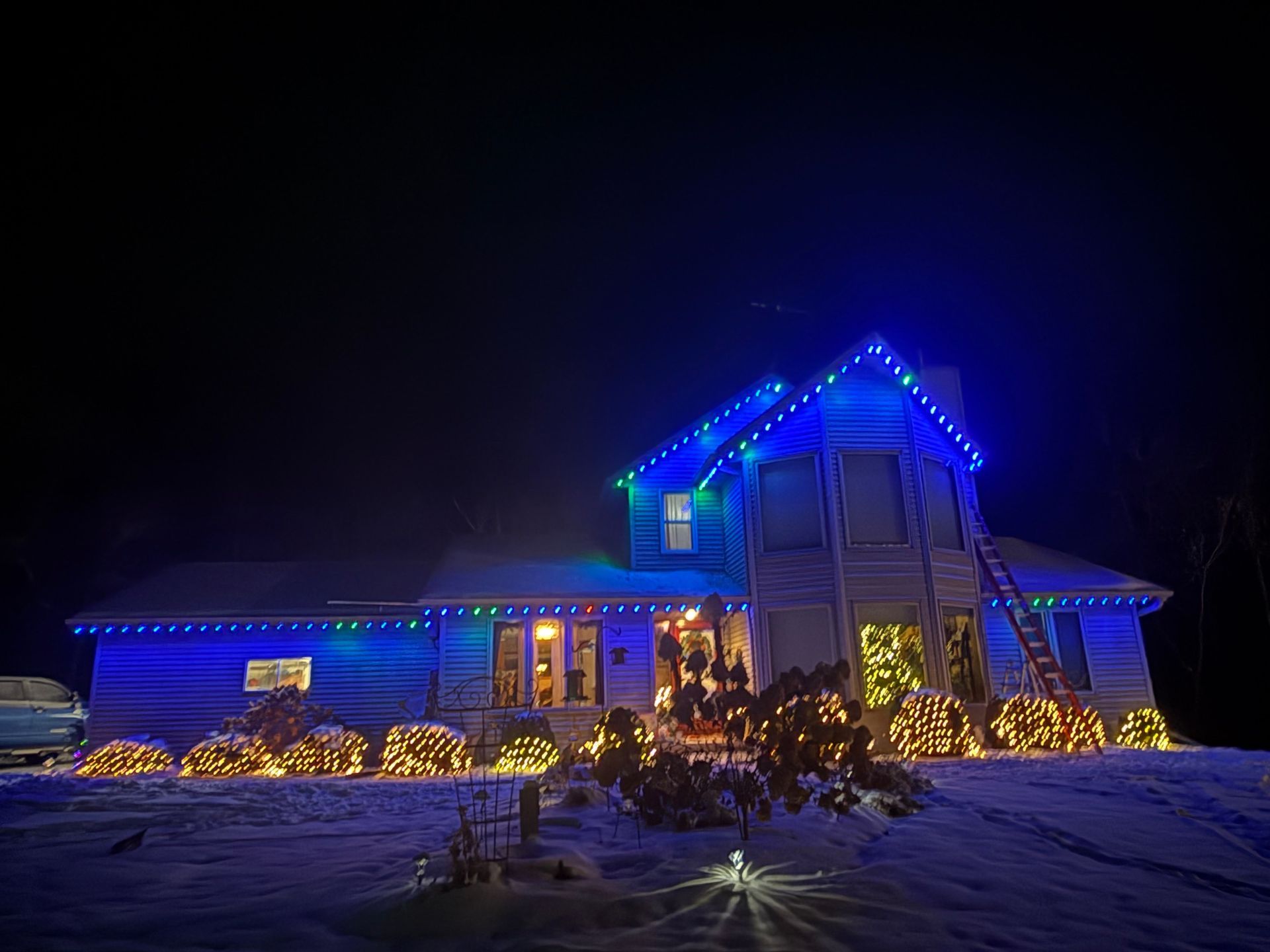 House at night with white lights outlining roof. Car parked on the left.