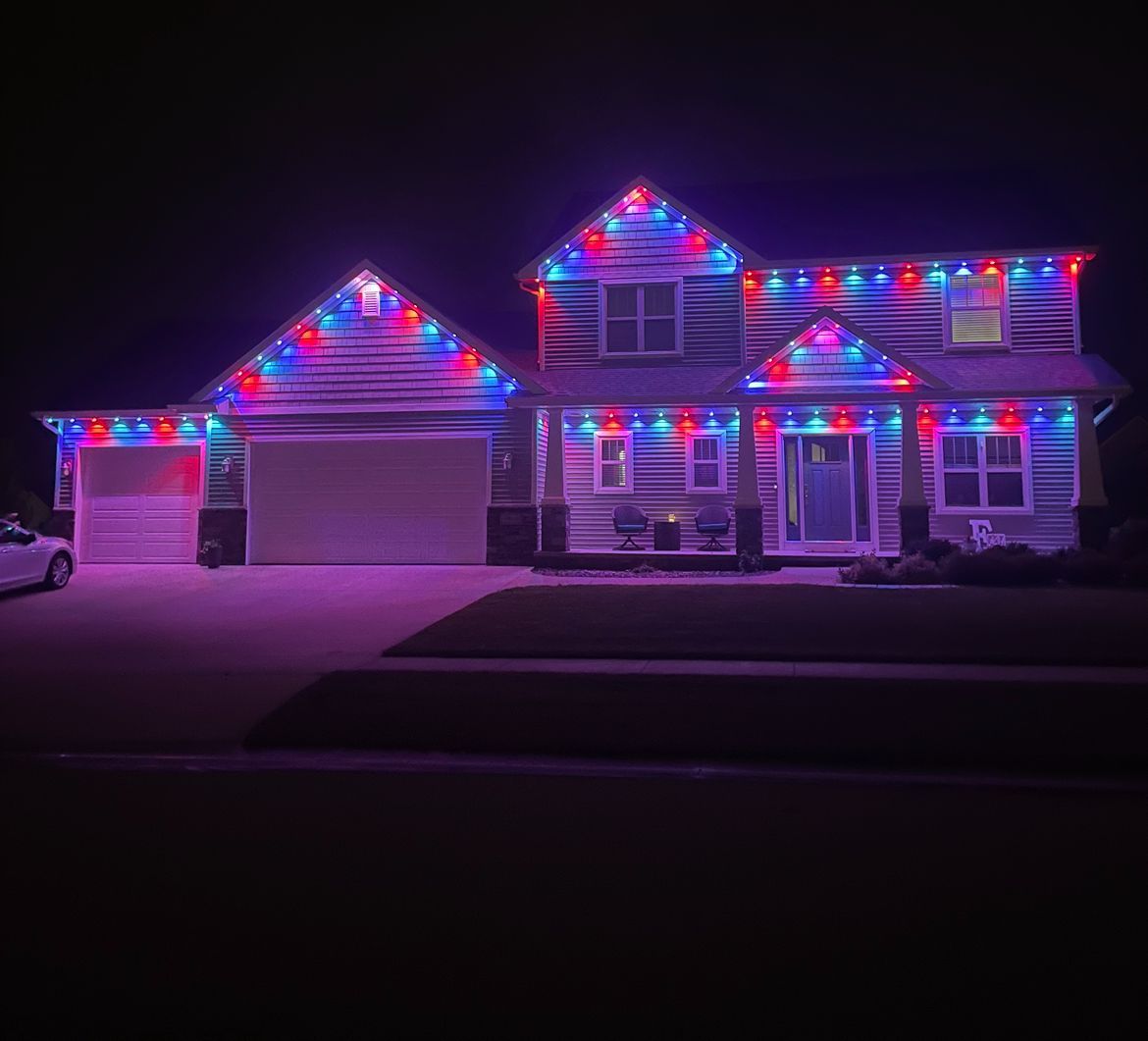 House decorated with red, white, and blue Christmas lights at night. Garage and porch light up the yard.