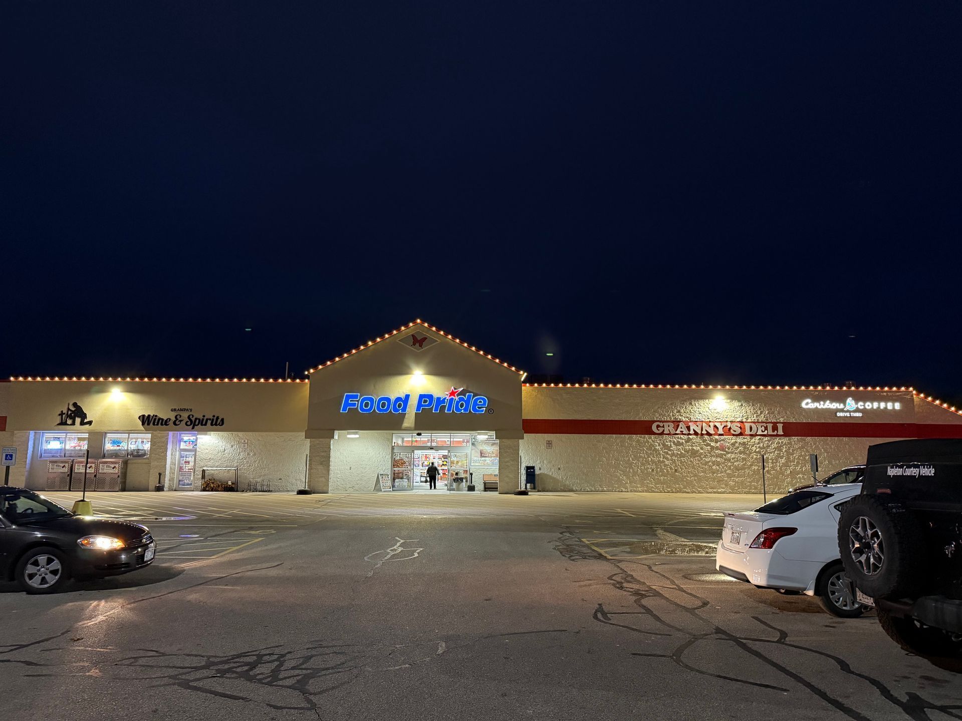 Exterior of a grocery store at night, lit up with lights. Cars parked in front. Dark sky above.