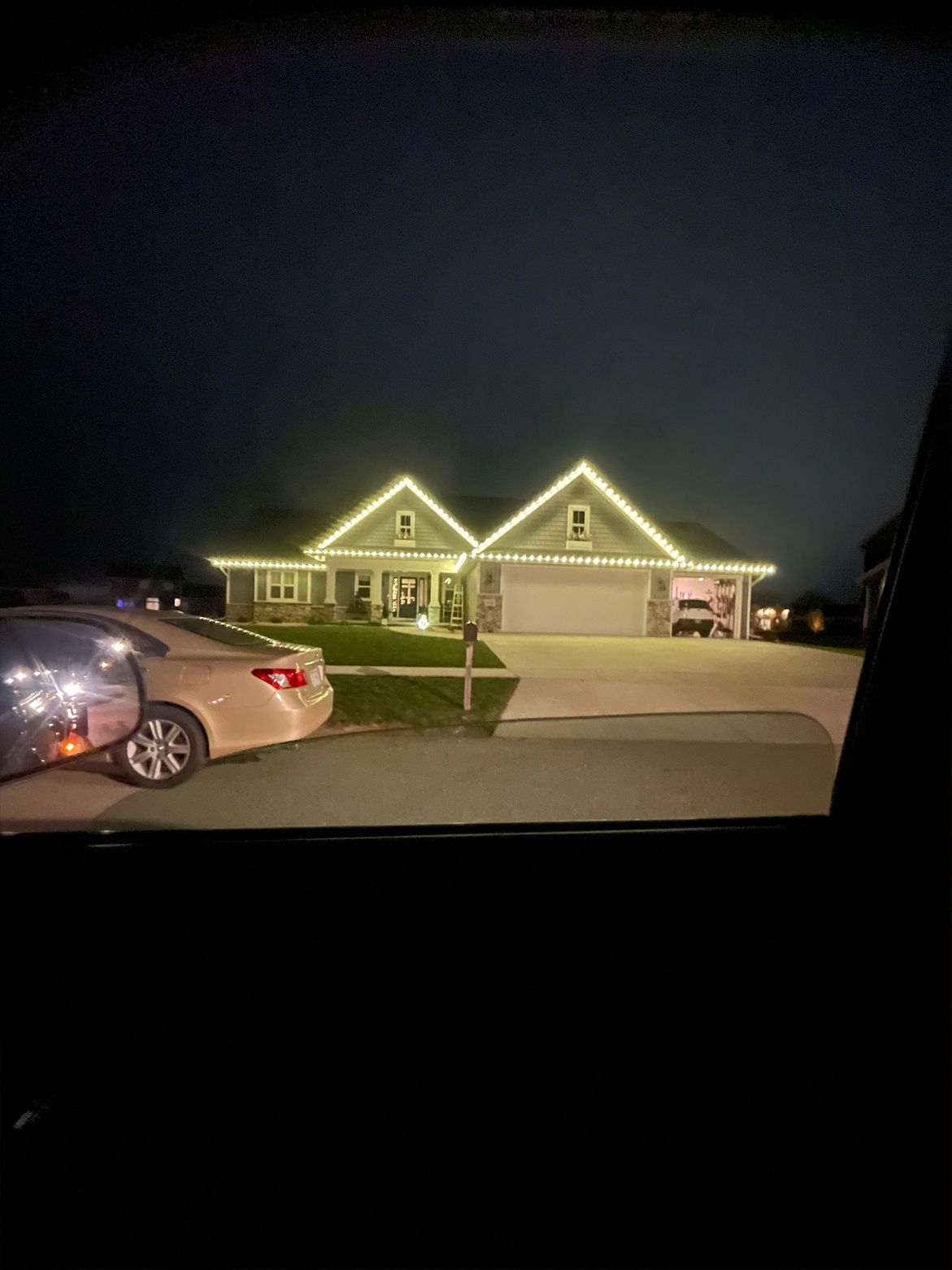 House at night with white lights outlining roof. Car parked on the left.