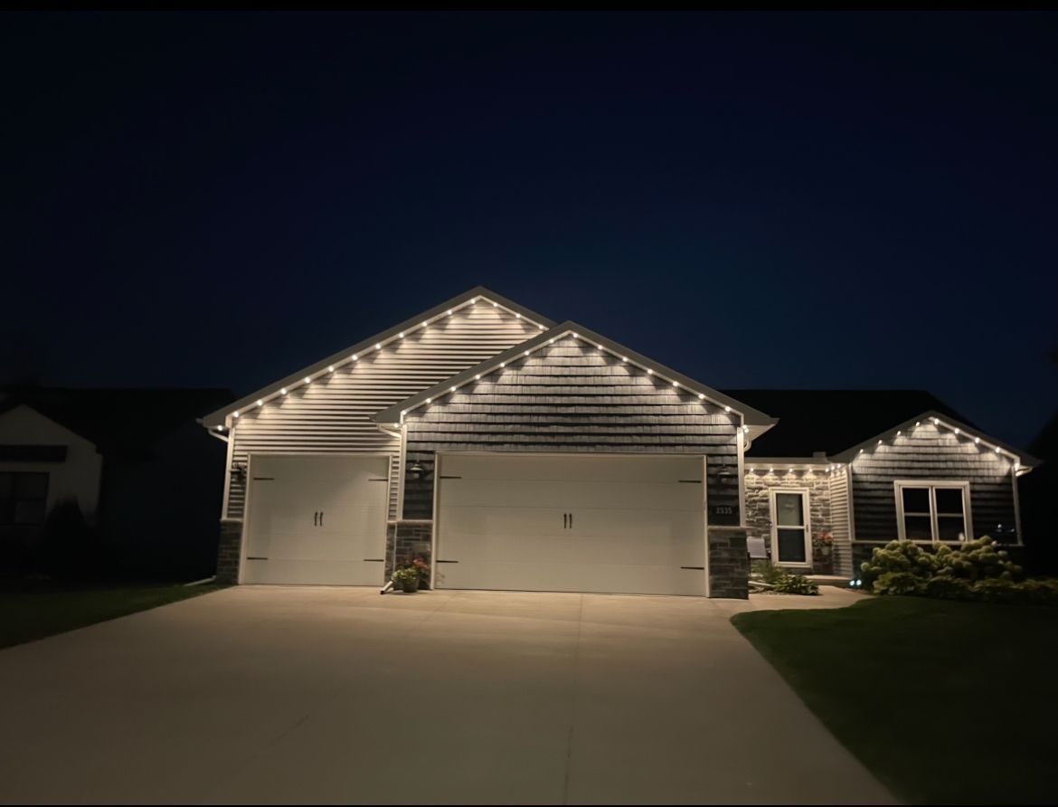 House at night, with white lights along the roofline. Two garage doors and driveway visible.