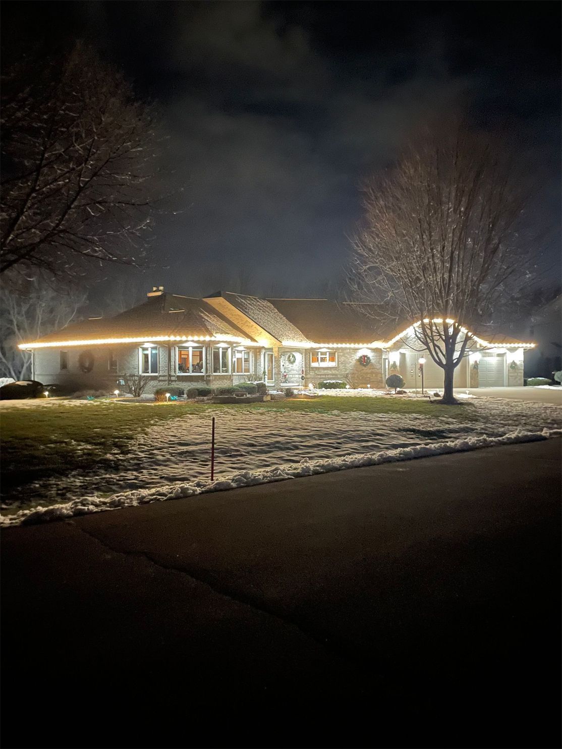 House at night with Christmas lights outlining roof and trees, snow-covered ground.
