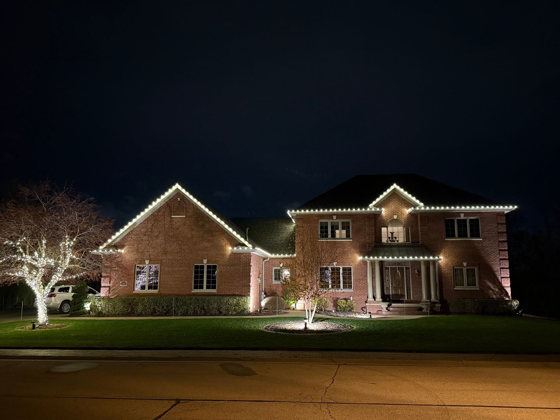 House decorated with white lights for the holidays. Lights outline the roof and trees in front.