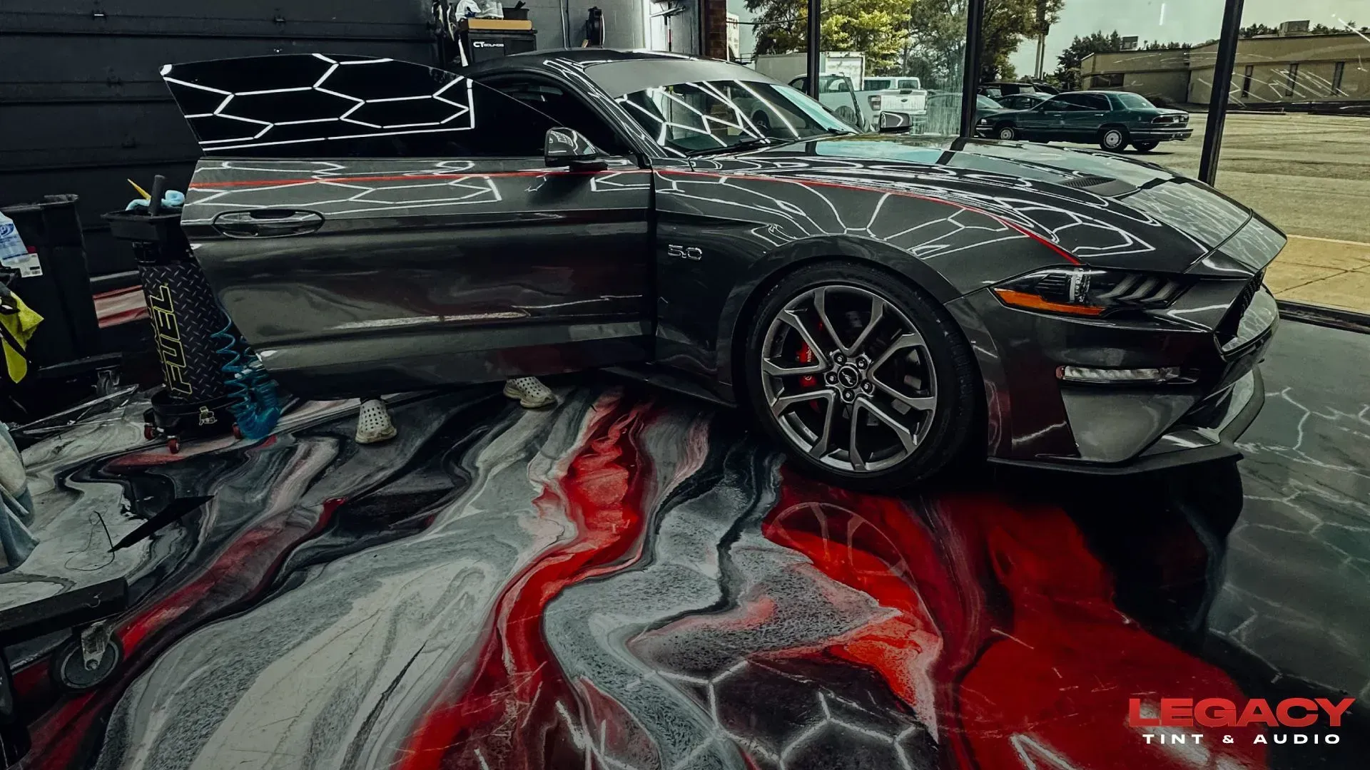 Gray sports car at an auto show, parked over a red-and-black marbled floor display.