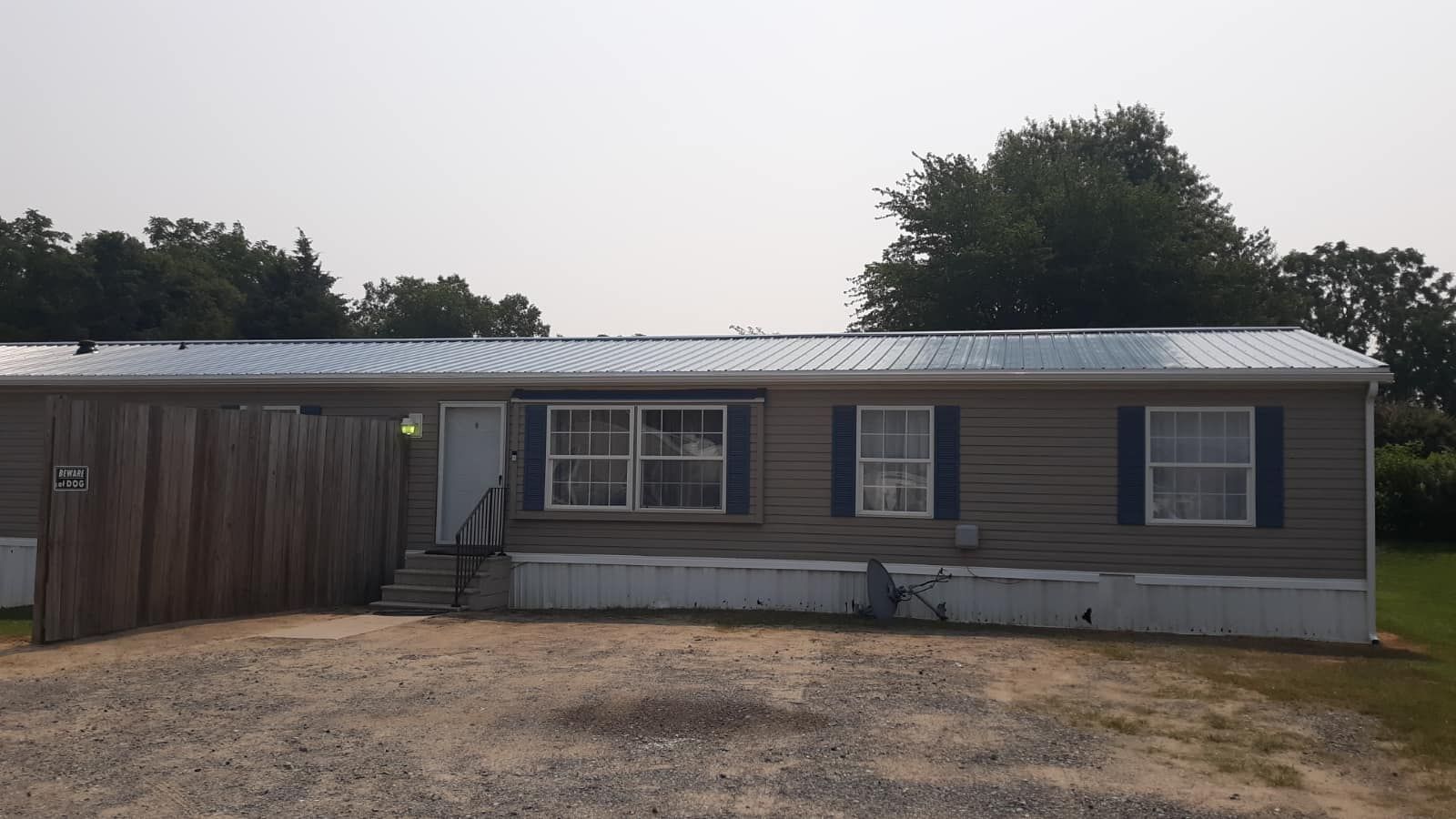 A mobile home with a metal roof and blue shutters is sitting in the middle of a dirt field.