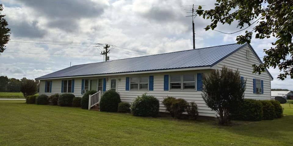 A white house with blue shutters and a metal roof is sitting on top of a lush green field.