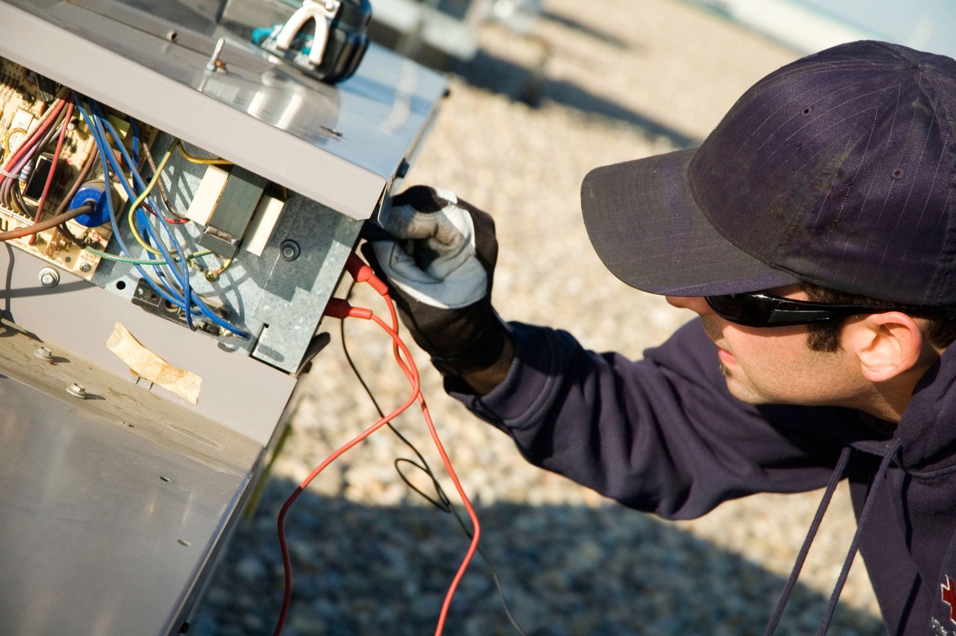 HVAC technician in cap and sunglasses tests wires inside AC unit on a rooftop.