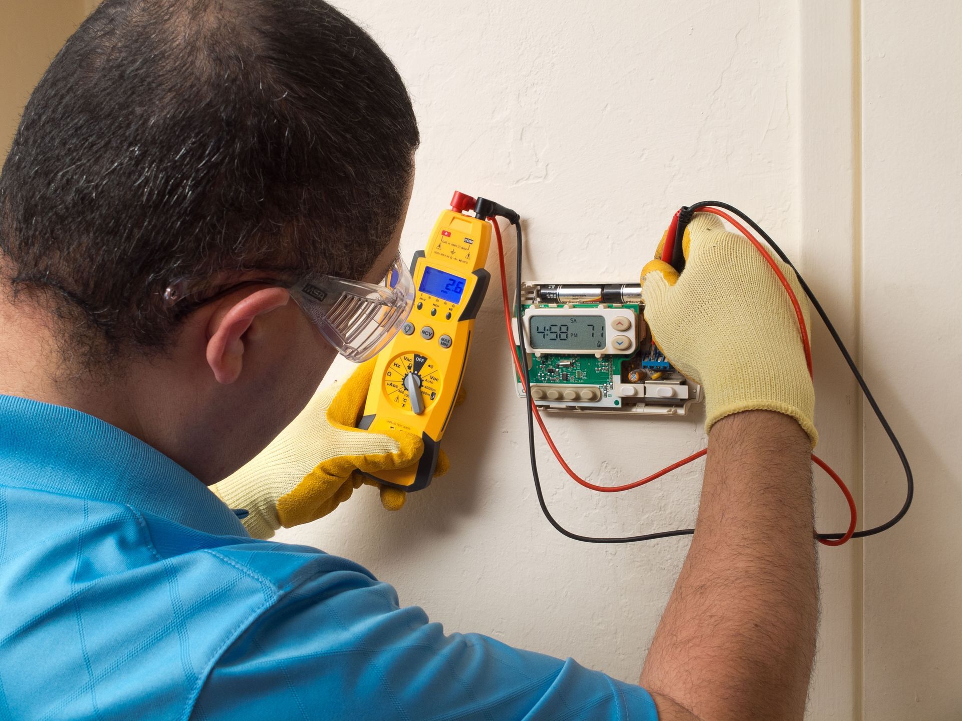 Person in safety glasses and gloves testing a thermostat with a multimeter on a wall.