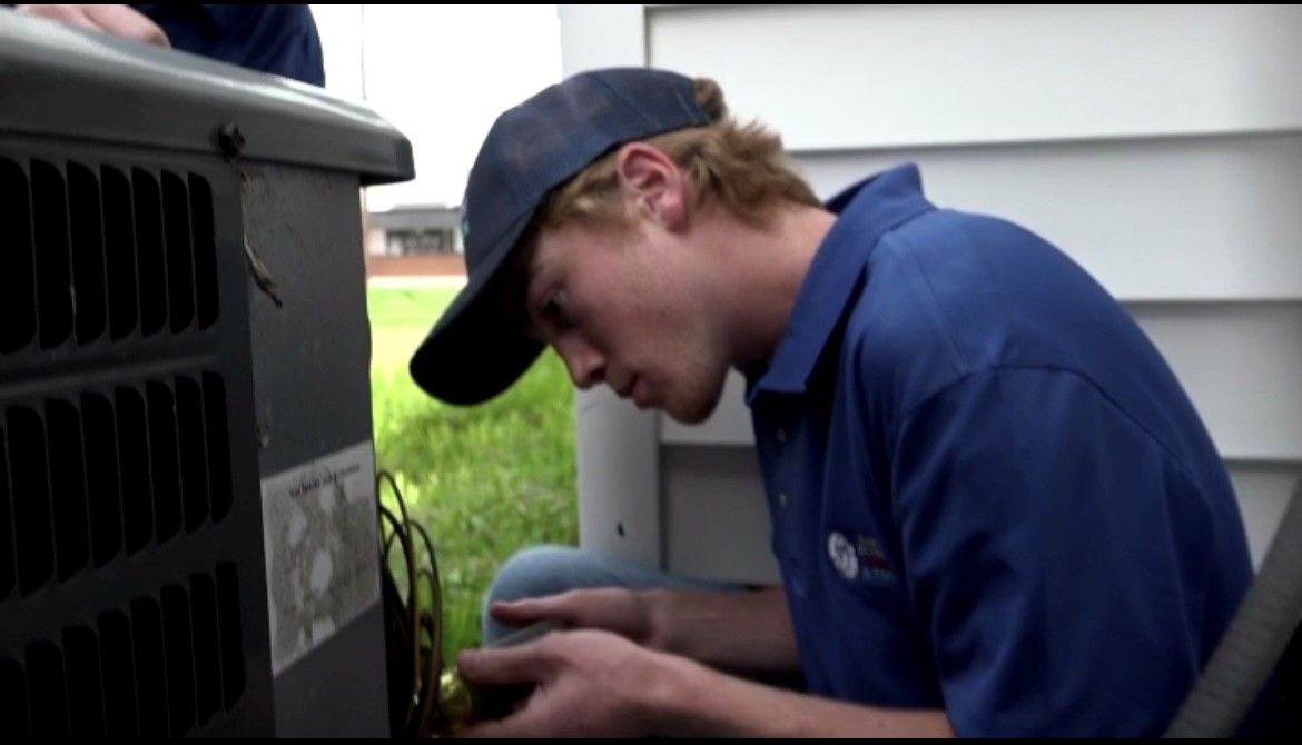 A technician in a blue shirt and cap inspects equipment beside a large outdoor unit.