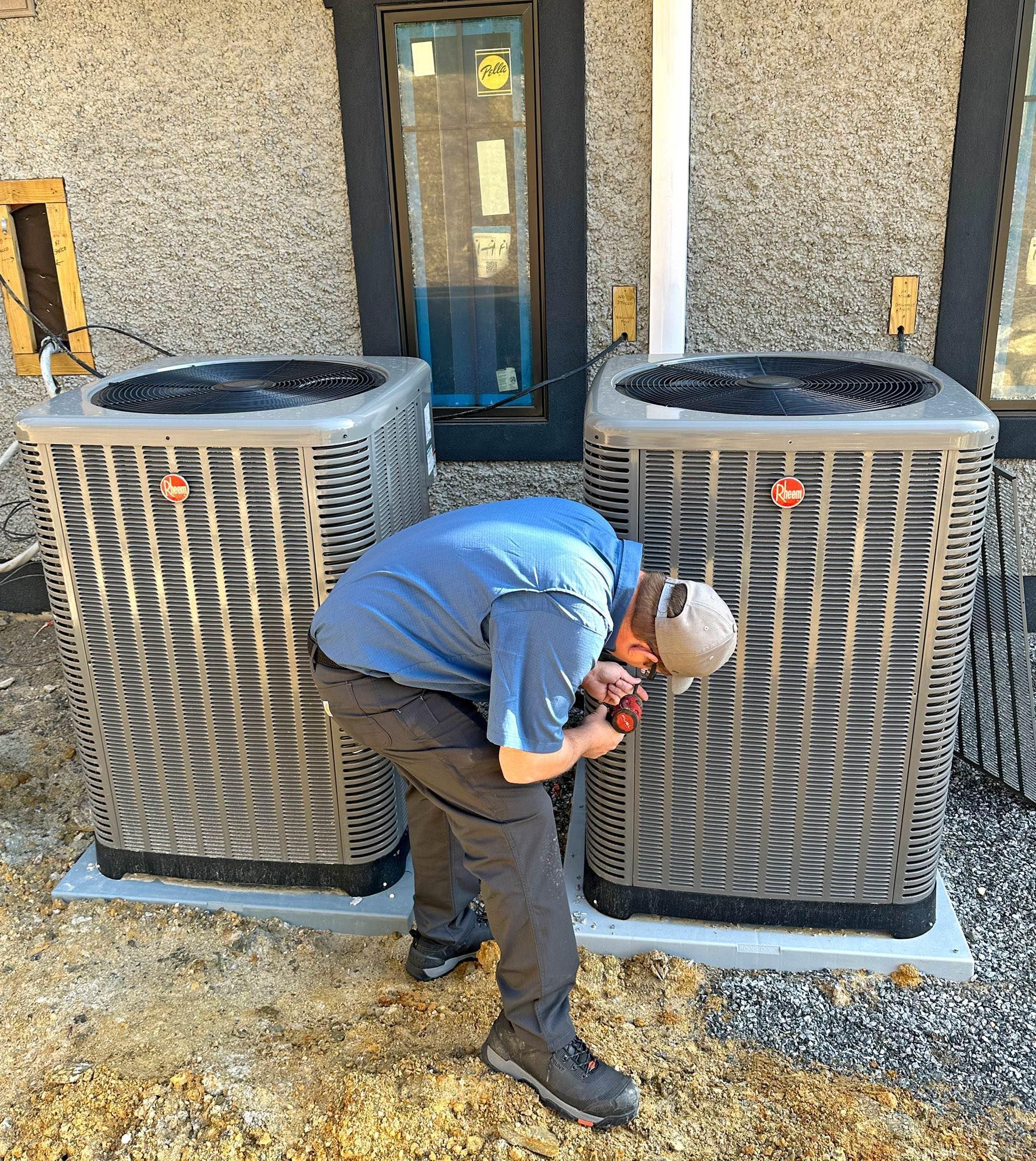 Man working on air conditioning units outside a building with gray stucco walls.