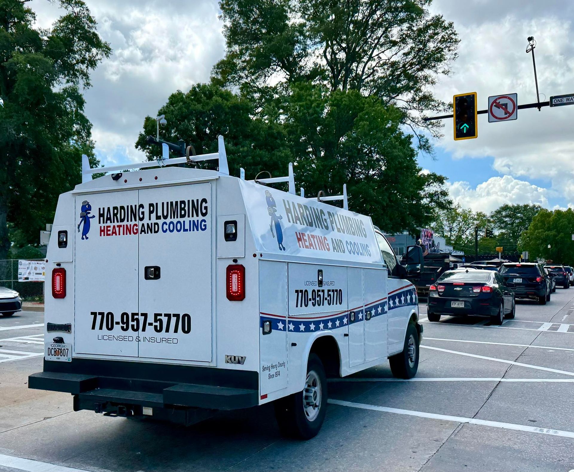 White Harding Plumbing van at a street intersection. Text & phone number on the side. Other vehicles and trees visible.