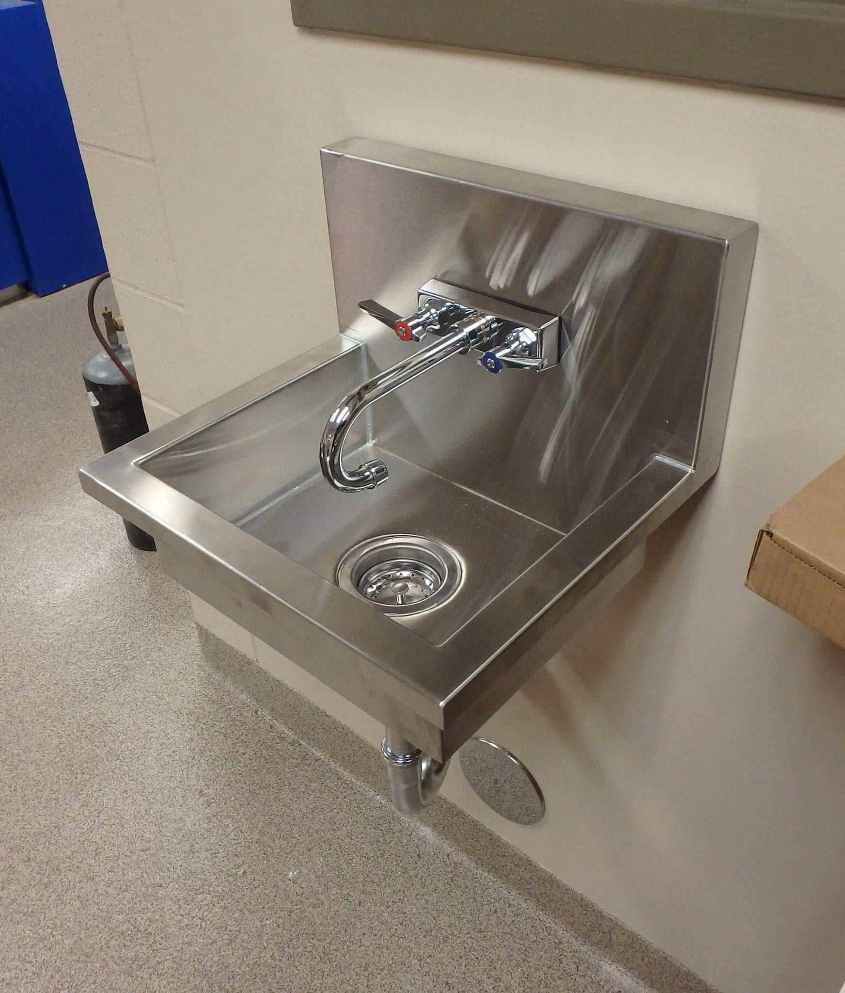 Stainless steel wall-mounted sink with faucet and drain, installed on a light-colored wall with speckled flooring.