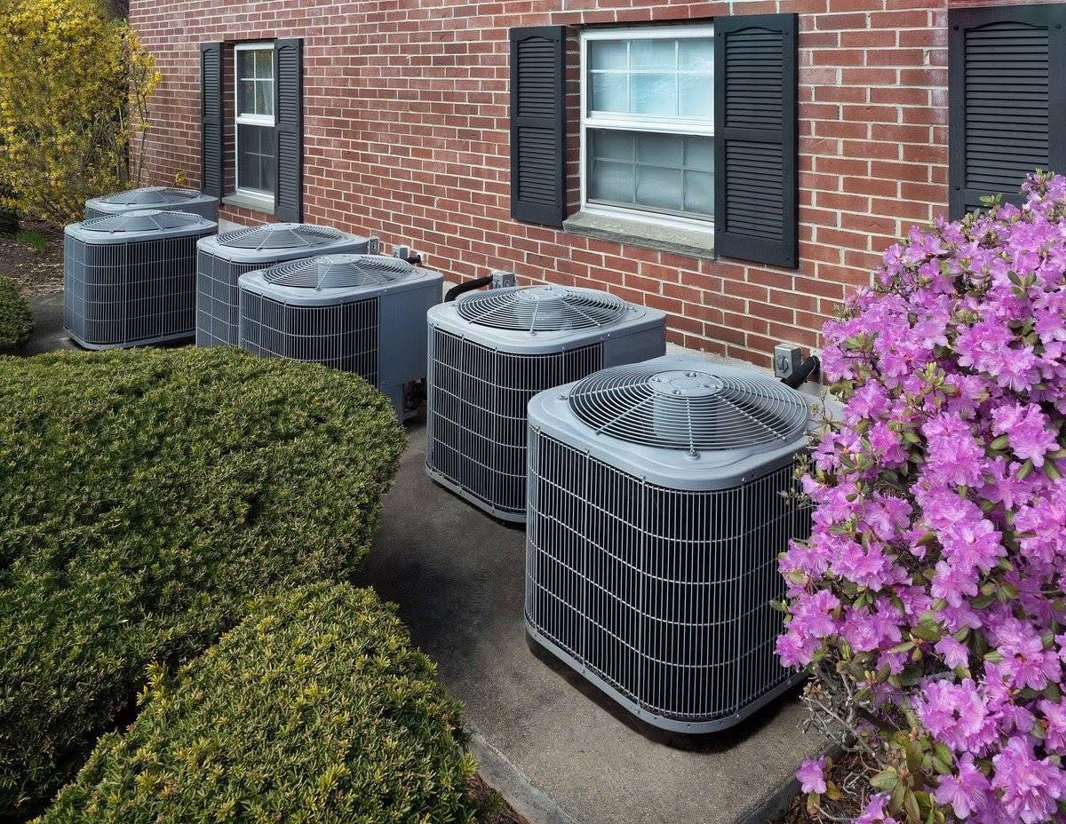 Several AC units next to a brick building and bushes, with azaleas blooming.