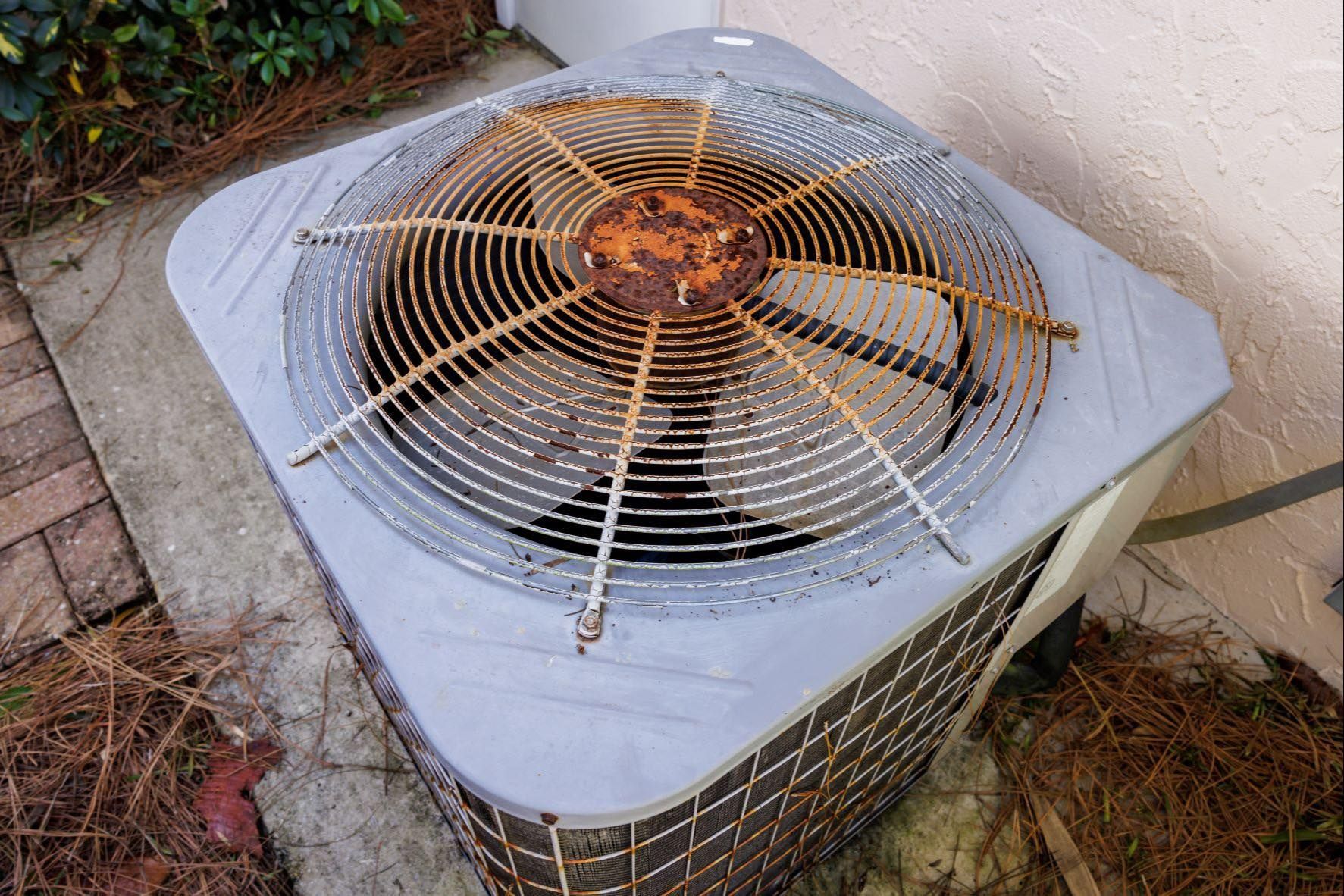 Rusted air conditioner unit with fan and protective grill, outdoors next to a wall.